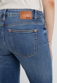 Siniset farkut, joissa on kapea muotoilu, varustettu ruskealla nahkatäggillä, jossa lukee "DRYKORN", sekä kuparivanteilla takataskuissa.