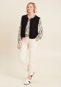 Schwarze Strickweste über einer gemusterten Bluse mit Paisleymuster, kombiniert mit hellen Hosen und weißen Sneakers mit schwarzen Akzenten.