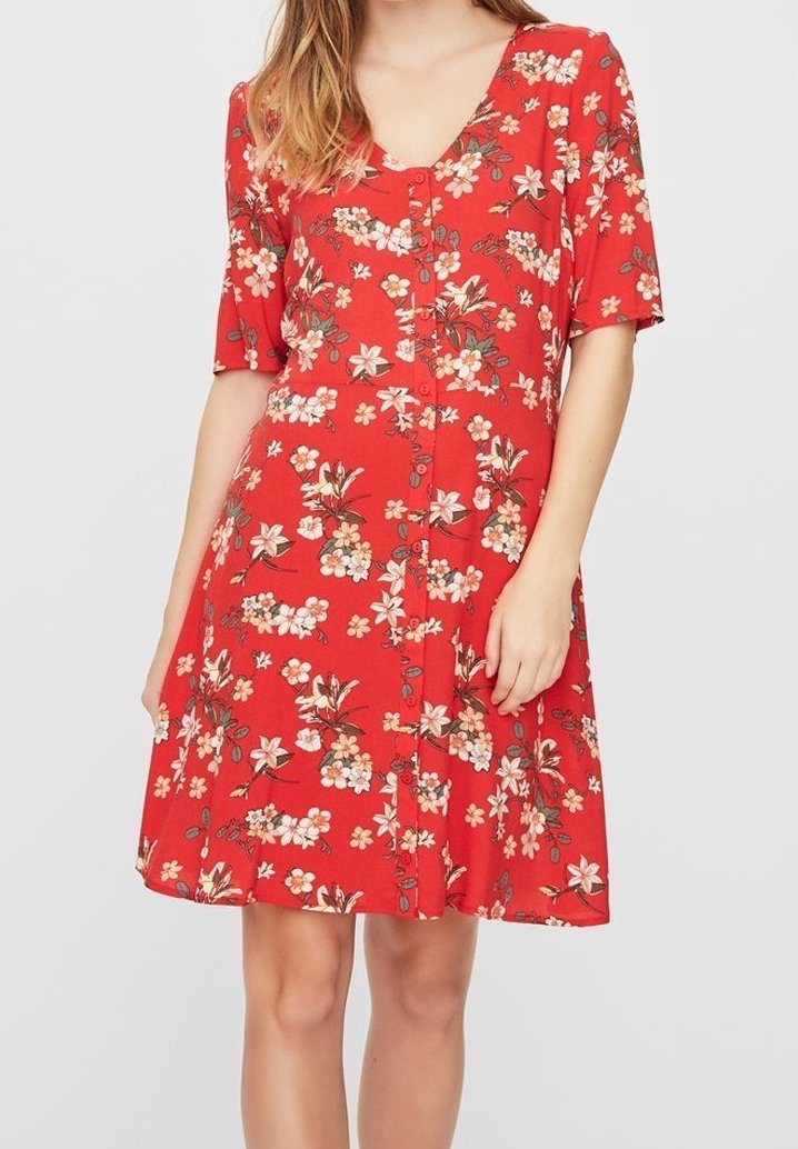 Femme portant une robe rouge à manches courtes avec des boutons sur le devant et des motifs floraux blancs et roses, se tenant devant un fond clair uni.