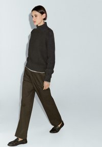 Maglione lavorato a maglia grigio scuro con colletto alto, texture a coste e dettagli sulle spalle, abbinato a pantaloni larghi marroni scuri e mocassini neri.