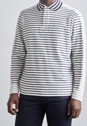 Hombre con una camisa polo blanca de manga larga con finas rayas horizontales azul marino y pantalones oscuros, de pie frente a un fondo liso.