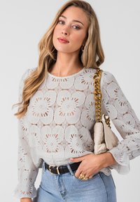 Light grey lace blouse with floral pattern, long sleeves, and ruffled cuffs. Paired with high-waisted blue jeans and a cream handbag.