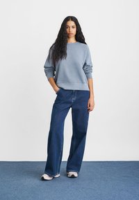 Sudadera de rizo gris con cuello redondo y mangas largas, combinada con pantalones vaqueros de pierna ancha de mezclilla azul de cintura alta. Zapatillas deportivas en tonos neutros.