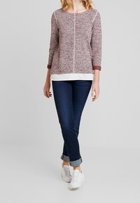 Suéter de punto burdeos con mangas tres cuartos, capa blanca debajo y puños en contraste. Combinado con jeans oscuros ajustados y zapatillas blancas.