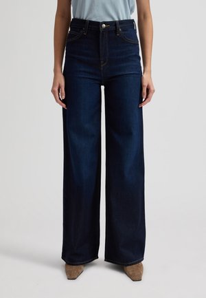 Άτομο που φοράει ψηλόμεσα, ανοιχτό μπλε τζιν με φαρδιά μπατζάκια, ανοιχτό γαλάζιο τοπ και μπεζ μυτερά παπούτσια, στέκεται μπροστά σε απλό φόντο.