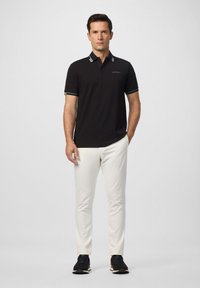 Muž stojaci čelne oblečený v čiernom polo tričku s krátkym rukávom, bielych nohaviciach a čiernych teniskách na jednofarebnom svetlom pozadí.