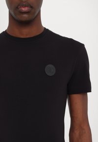 Tricou din bumbac negru cu mâneci scurte, guler rotund și o etichetă din cauciuc pe pieptul stâng. Textură netedă și design ajustat.