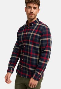 Rood en marineblauw geruit overhemd met knopen, gemaakt van flanel, met lange mouwen, een kraag en bruine knopen. Draagt het over olijfgroene broek.