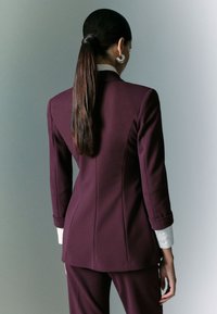 Blazer sagomato viola scuro con maniche lunghe e vestibilità strutturata. Presenta un polsino di camicia bianca visibile sulla manica. Tessuto dalla trama liscia.