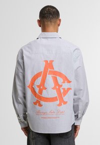 Stribet langærmet skjorte i lysegrå med et orange oversized logo-design på ryggen, har en knappet manchet og afslappet pasform.
