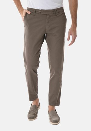 Uomo con pantaloni marroni aderenti e scarpe slip-on grigie, in piedi con una mano in tasca su sfondo bianco.