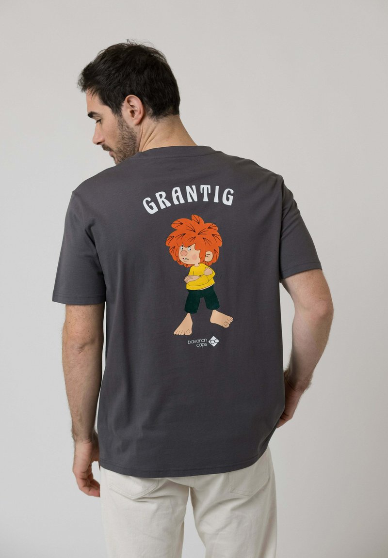 Man with dark hair wears a dark gray T-shirt featuring a cartoon red-haired child crossing arms with "GRANTIG" text above on the back.