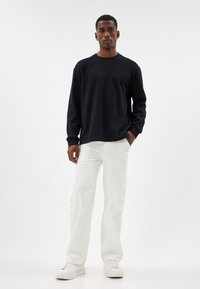 Svart långärmad tröja i texturerat tyg, kombinerad med vita cargobyxor. Modellen bär också vita sneakers och står på en ljus bakgrund.