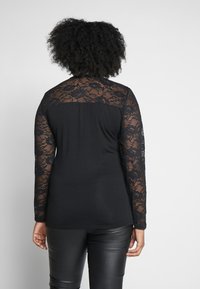 Haut noir à manches longues avec des détails en dentelle sur les manches, présentant un dos uni et une texture lisse. Assorti à un pantalon noir ajusté.