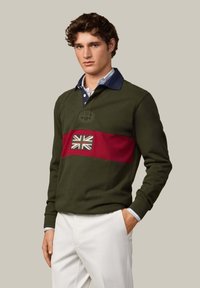 Zielona bawełniana koszulka polo z bordowym poziomym paskiem ozdobionym haftowanym Union Jack. Ma granatowy kołnierzyk i jest zestawiona z białymi spodniami.