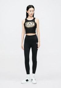 Black crop top with gold logo on front, paired with high-waisted black leggings. Model wears white sneakers with contrasting black accents.