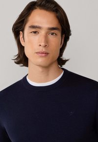 Marineblauwe trui met een ronde hals, geribbelde manchetten en een subtiel logodetail op de borst. Zachte textuur, getailleerd ontwerp.