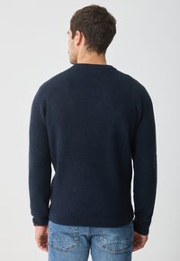 Dunkelblaues Strickoberteil mit Rundhalsausschnitt, gerippten Bündchen und Saum. Weiche Textur mit klassischer Passform, getragen über blauen Jeans.