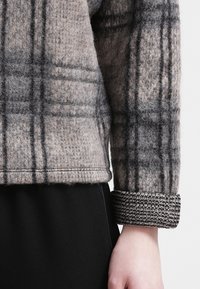 Gros plan d'une personne portant une veste en laine grise à carreaux avec un poignet texturé sur un pantalon noir, montrant le bras et la main.
