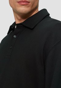 Schwarzes Poloshirt mit geripptem Kragen, Dreiknopf-Blende und strukturiertem Stoff. Nahaufnahme des Kragens und des Schulterbereichs.