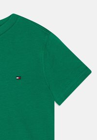 T-shirt verde a maniche corte con collo rotondo e piccolo logo rettangolare rosso, bianco e blu sul petto, con cuciture visibili sulle maniche e sulle spalle.