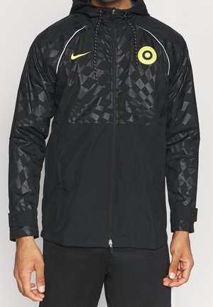 Veste à capuche noire zippée avec tissu brillant à motifs sur les manches et la poitrine, logo jaune Nike et emblème circulaire à l'avant.
