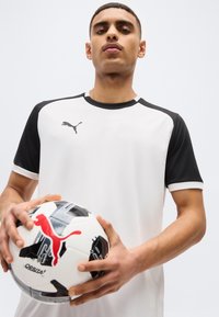 Homme en tenue de sport Puma blanche et noire tenant un ballon de football de la marque Puma sur un fond clair uni.