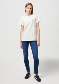 T-shirt en coton beige clair à col rond avec un petit logo navy sur la poitrine, associé à un jean skinny bleu foncé et des baskets noires.
