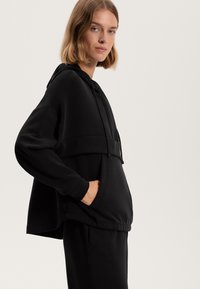 Felpa nera con cappuccio dalla vestibilità comoda, dotata di una tasca frontale e coulisse visibili. Realizzata in tessuto morbido, con una texture opaca.