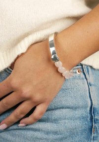 Sölj cuff-armband med ett hjärtdetalj, parat med ett pärlband i ljus rosa rosenkvarts. Bärs på handleden mot denim.