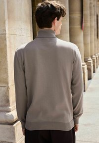 Taupe gebreide trui met een hoge kraag en geribde manchetten. Zachte textuur, slim fit ontwerp, zichtbare stiksels bij de halslijn en de tailleband.