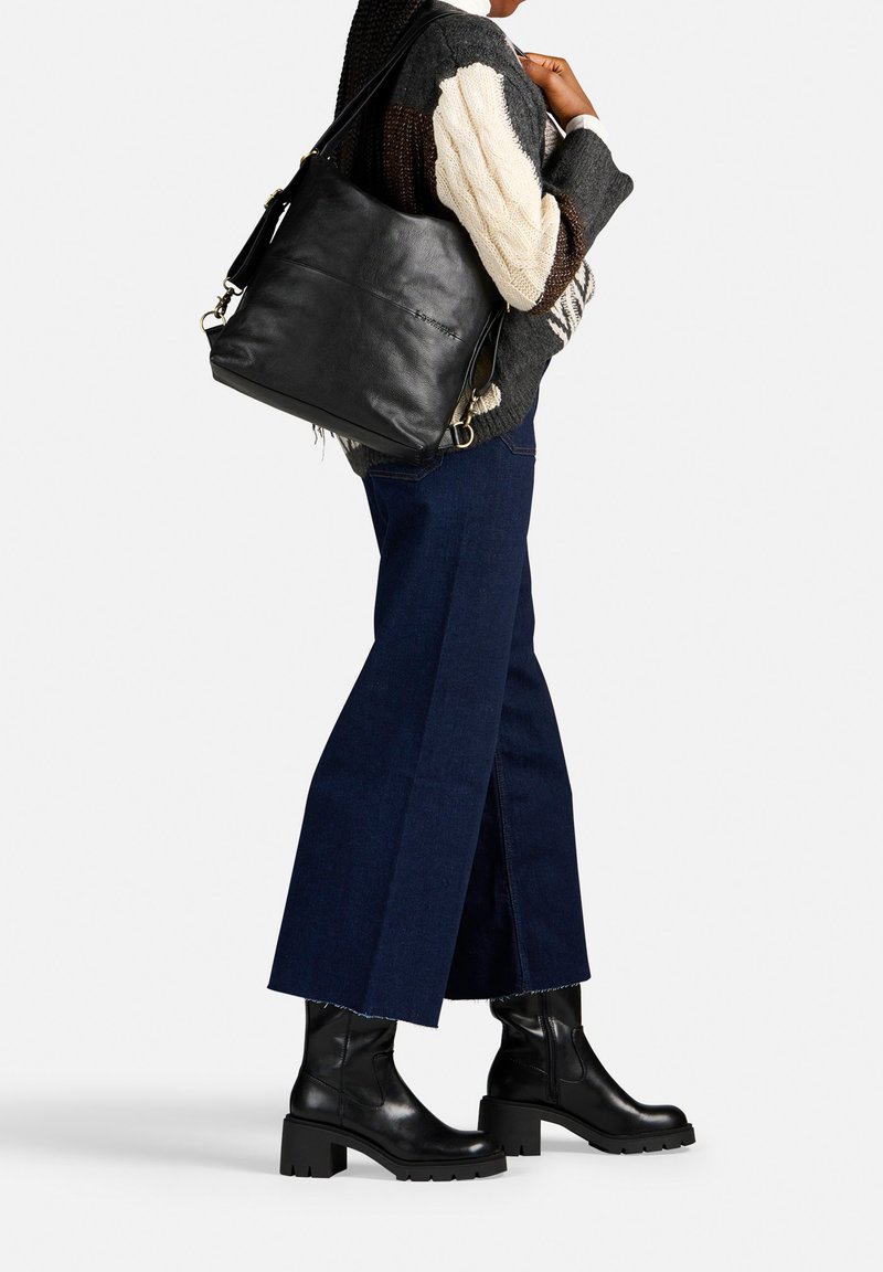 Borsa a tracolla nera in pelle con hardware dorato, indossata sopra un maglione grigio e bianco, abbinata a un paio di jeans a gamba larga blu scuro e stivaletti neri.