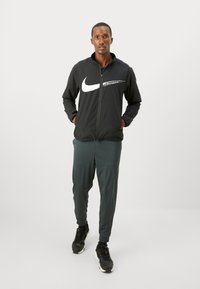 Sort zip-up jakke med hvidt Nike-logo, parret med mørkegrønne tapered bukser og sorte sneakers. Minimalistisk design og atletisk pasform.