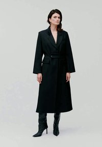 Casaco preto longo com um ajuste sob medida, lapelas largas, fecho com um botão único e cinto à condizer. Usado com botas altas pretas de salto.
