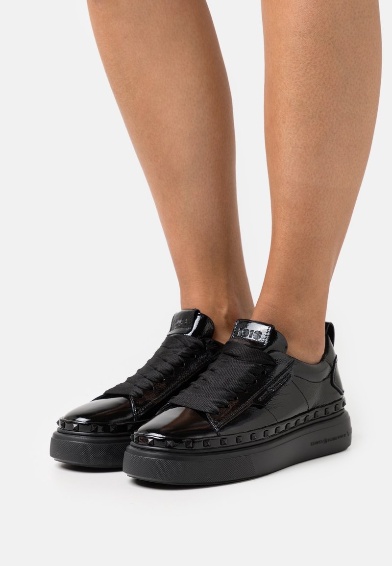 Baskets basses noires brillantes avec semelles épaisses et lacets larges portées sur des jambes nues sur un fond uni.