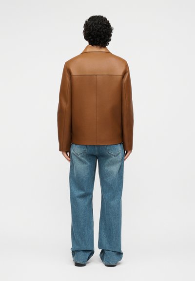 Persona con capelli ricci che indossa una giacca di pelle marrone e jeans blu larghi, in piedi di spalle contro uno sfondo semplice.