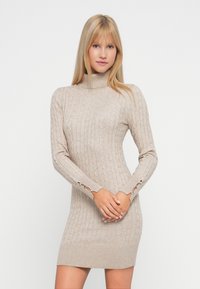Mujer rubia con un vestido de punto de tortuga beige y detalle de botones en las mangas, de pie frente a un fondo liso.