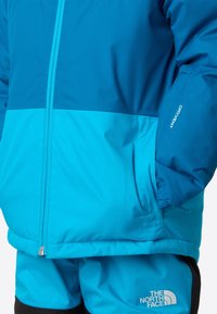 Nepremokavá bunda v žiarivej modrej a tyrkysovej látke, s predným zipsom, vreckami a logom. Ľahký a priedušný dizajn.