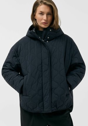 Kobieta ubrana w czarną pikowaną kurtkę puchową z wysokim kołnierzem, z rękami w kieszeniach, pozująca na jednolitym jasnym tle.