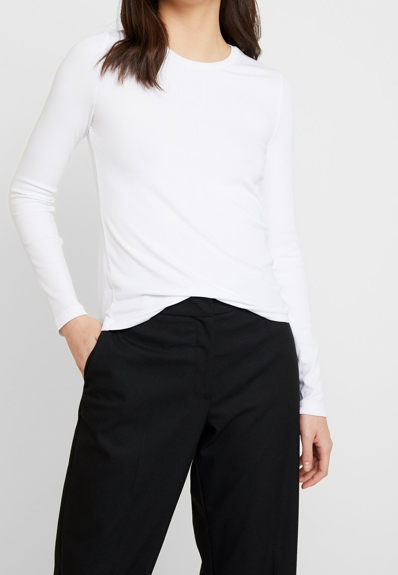 Haut blanc à manches longues en maille côtelée avec un col rond, associé à un pantalon noir taille haute. Texture lisse ; sans motifs ni accents visibles.