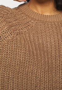 Brauner Strickpullover mit einem gerippten Kragen und strukturiertem Muster, der eine lockere Webart und deutlich sichtbare Nähte aufweist.