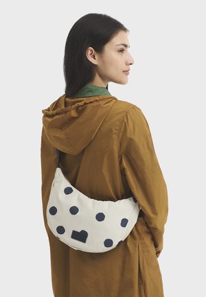 Femme portant une veste à capuche marron avec un sac blanc en forme de croissant orné de gros pois noirs, pendu à son épaule.