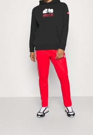 Mężczyzna w czarnej bluzie z kapturem Adidas Adventure, jaskrawoczerwonych spodniach dresowych Adidas z białymi paskami oraz czarno-białych trampkach, stojący.