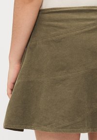 Olijfgroene corduroy rok met een uitlopende zoom, voorzien van horizontale naaddetails en een soepele tailleband. De textuur is ribbelig en zacht.