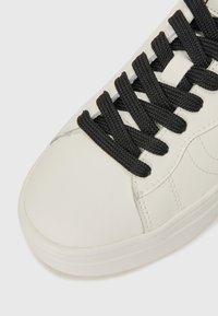 Sneaker bianco in pelle con texture liscia, caratterizzato da lacci intrecciati neri e dettagli di cucitura sottili lungo i lati. Design con punta rotonda.