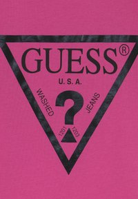 Rozā kokvilnas t-krekls ar lielu melnu trīsstūra logotipu, kurā ir teksts "GUESS", "U.S.A." un "WASHED JEANS", kā arī jautājuma zīme un skaitļi 1201, 1203.