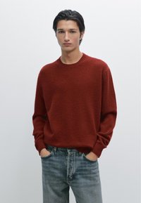 Röd stickad tröja med rund halsringning och ribbad manschett, i kombination med urtvättade blå jeans. Texturen verkar mjuk och något lurvig.