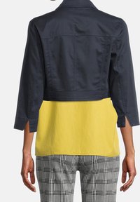 Kvinna som bär en kort mörk marinblå jacka över en gul topp och svartvit rutiga byxor, sedd bakifrån.