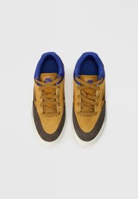 Adidași din suede maro și galben, cu accente albastre, prevăzuți cu șireturi, guler moale și o talpă din cauciuc alb. Designul include detalii cu logo.