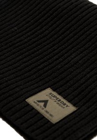 Sort ribbet strikket beanie med et kvadratisk vævet mærke, der viser "SUPERDRY" og et triangel logo. Materialet ser blødt og strækbart ud.